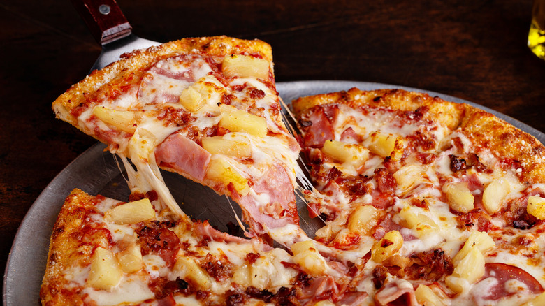 Someone uses a spatula to lift a slice of Hawaiian pizza