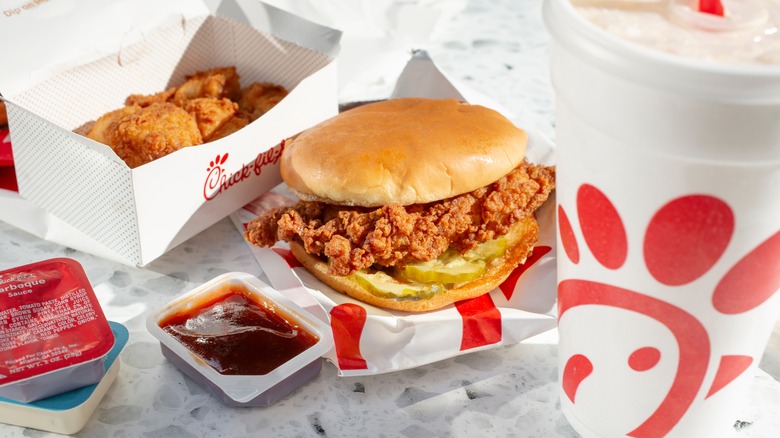 Chick-fil-A chicken sandwich, box with nuggets, and a drink on a white table with an open red sauce cup