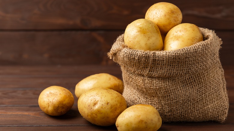 Burlap sack filled with potatoes, with a few potatoes outside of it on a dark wooden surface
